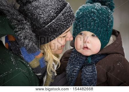 Portrait of young mother and little toddler boy in the winter forest. Family enjoying winter. Child and woman watching falling snow outdoors. Winter Christmas and lifestyle concept.