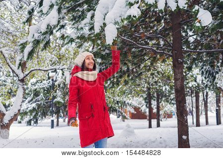 A girl holding a branch with snow a young girl walks through the park and enjoying the first snow winter day. Red coat and gloves knit cap. The snow is showered from trees.