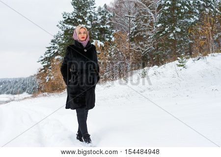 The girl walks on a snow trail along the shore and woods. Snowy autumn day winter is coming. Warm clothing a coat and a scarf on her head. Russian beauty.