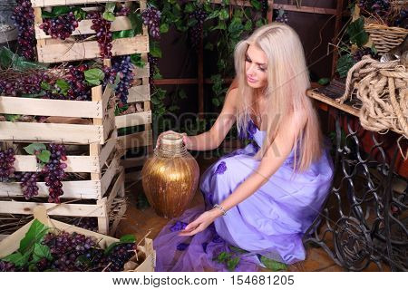 Pretty blonde woman in dress poses with jug among boxes of grape in studio