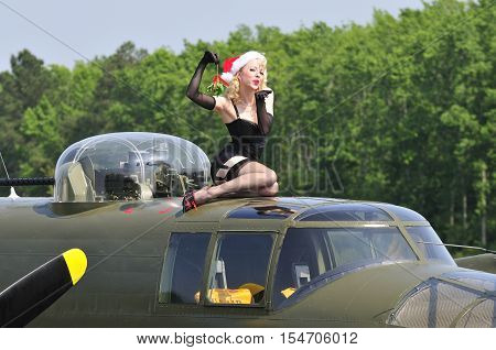 Sexy Christmas pinup holding up a mistletoe and blowing a kiss while sitting on top of a WWII bomber aircraft