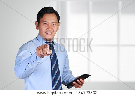 Photo image of a handsome attractive successful young Asian businessman smiling and pointing finger to you towards front camera while holding a tablet