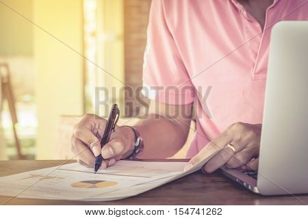 Business man hand holding pen point graph chart documen and use computer laptop in office.