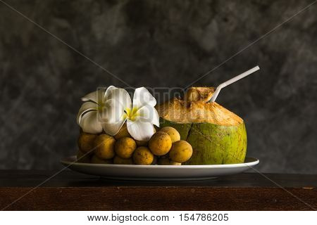 Coconut with southern langsat ornamated with plumeria in white dish on wooden table.