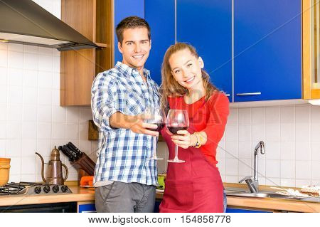 Young couple cheers with red vine in the kitchen at home - Happy boyfriend and girlfriend celebrating relationship drinking together inside the house - Concept of joyful moment love and happiness