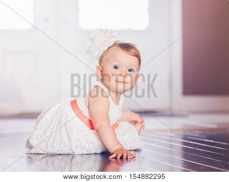 Portrait of cute adorable blonde Caucasian smiling baby child girl with blue eyes in white dress with red bow sitting on floor indoors looking in camera dreaming fairy tale sun light from above behind