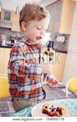 Portrait of one happy kid boy eating breakfast waffles with fruits with fork in sunny kitchen early morning showing his tongue