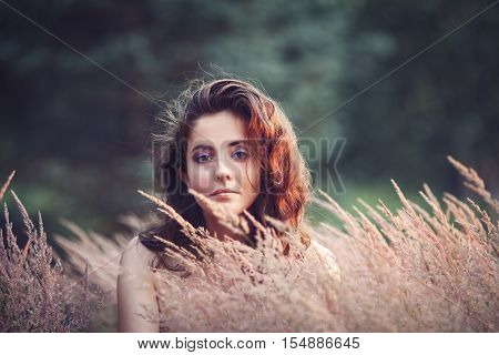 Portrait of beautiful innocent Caucasian adult girl woman with long dark red brown hair hazel eyes in park field outside on sunset in golden hour looking in camera