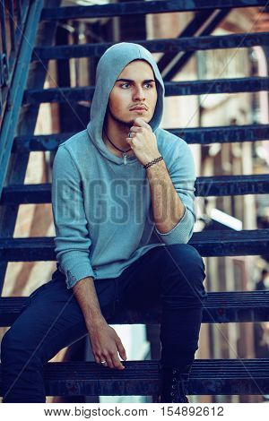 Conceptual art portrait of beautiful handsome pensive young middle east brunette man with blue eyes beard wearing blue hoodie sitting on staircase outside in street looking away