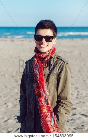 Portrait of happy smiling beautiful Caucasian white brunette woman with short hair in green jacket red scarf and sunglasses looking in camera standing on sea ocean beach at sunset