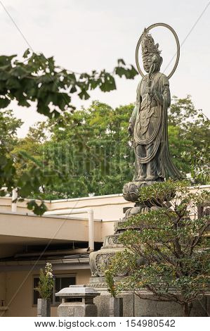 Tokyo Japan - September 26 2016: Stone statue of Bodhisattva Quan Yin standing on lotus pedestal in garden at Senso-ji Buddhist Temple. Large halo. Green foliage. Gray sky.