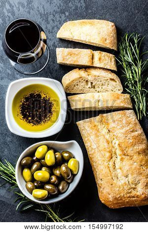 Bread ciabatta, olives, arugula, olive pepper oil, olives, rurosemary and glass of red wine on stone slate black background. Top view copy space