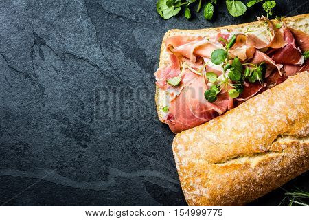 Ciabatta sandwich with jamon ham serrano paleta iberica, arugula, rosemary on stone slate black background. Top view