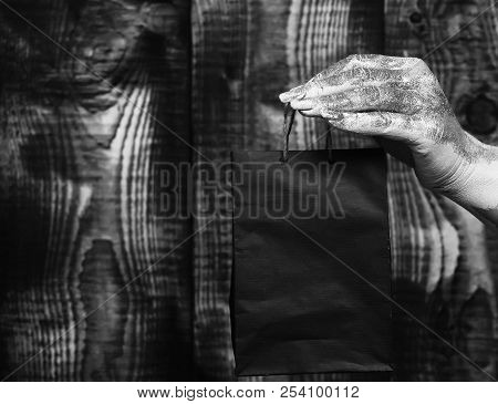 Female Hand Smeared In Golden Paint Or Glister Holding Black Selective Focus Package Or Bag On Brown