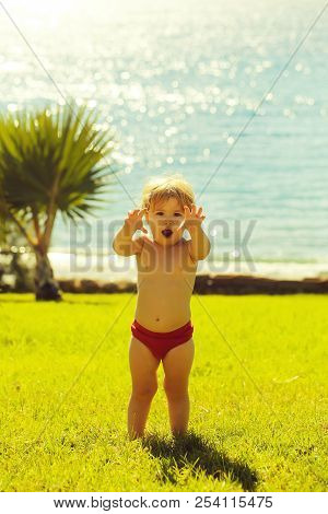 Cute Baby Boy With Blond Hair In Red Trunks Makes Frightening Attacking Gesture Outdoors On Idyllic,