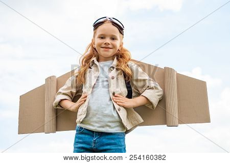 Low Angle View Of Child With Paper Plane Wings On Back And Protective Eyeglasses On Head Against Blu