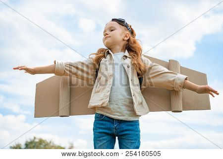 Low Angle View Of Child With Paper Plane Wings And Protective Eyeglasses With Outstretched Arms Agai