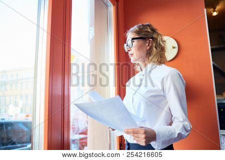 Elegant pensive woman in glasses holding papers and looking away in window contemplating on work