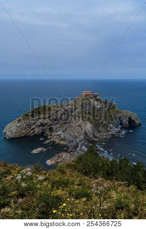 Vertical Shot Containing Some Foliage At The Front, The Whole Dragonstone-gaztelugatxe Island At The