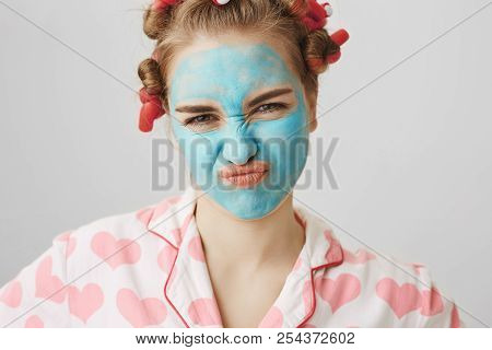 Close-up Portrait Of Nasty Meany Girl In Facial Mask And Hair-curlers, Wearing Pyjamas And Squinting