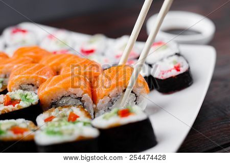 Eating Sushi. Food Photo, Japanese Restaurant, Dinner. Pair Of Chopsticks Taking Sushi Roll From Pla
