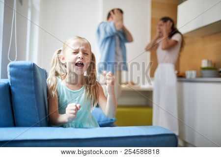 Little capricious girl expressing her displeasure by loud voice and hitting armchair with shocked parents on background