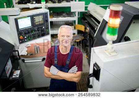 Portrait of confident mature operator standing in overall with arms crossed standing at workplace among computer machines
