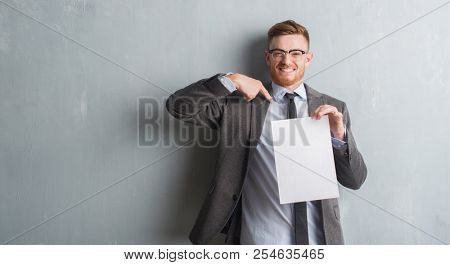 Young redhead  business man over grey grunge wall holding blank paper contract with surprise face pointing finger to himself