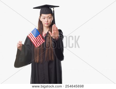 Young Chinese woman wearing graduate uniform holding United states of america flag with open hand doing stop sign with serious and confident expression, defense gesture