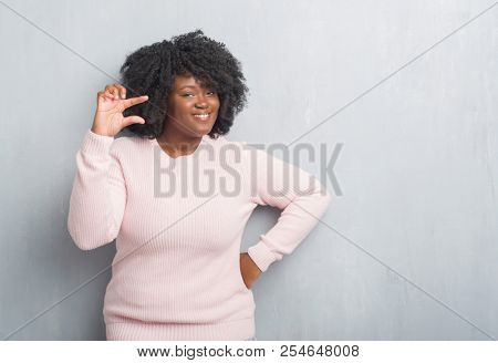 Young african american plus size woman over grey grunge wall wearing winter sweater smiling and confident gesturing with hand doing size sign with fingers while looking and the camera. Measure.