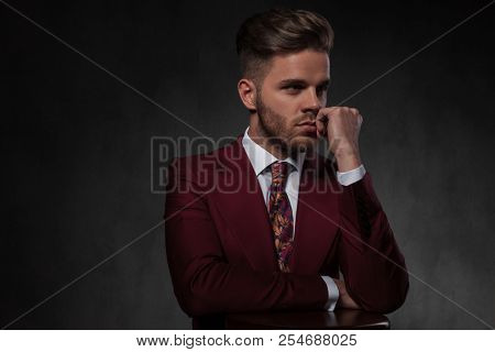 portrait of pensive elegant man resting his hands on wooden chair and looking to side while standing on grey wallpaper background