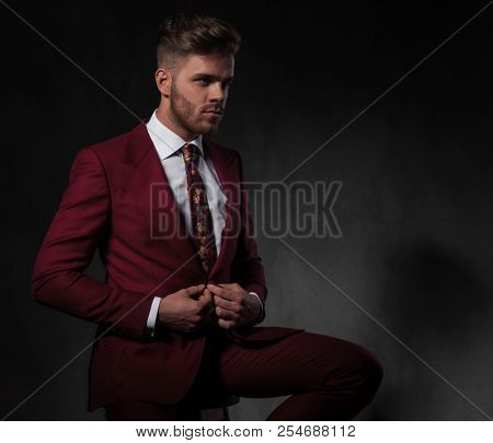 seated man buttons his red suit while looking to side on grey wallpaper background