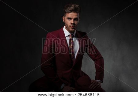 handsome young man wearing a grena colored suit sitting on grey wallpaper background while resting his hand on his thigh
