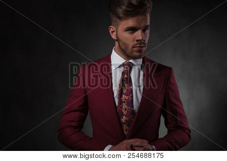 portrait of young man in red suit holding palms together while looking to side and standing on grey wallpaper background