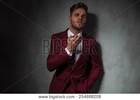 portrait of seductive man in grena suit sending an air kiss while standing with a hand in pocket and leaning against a grey wall