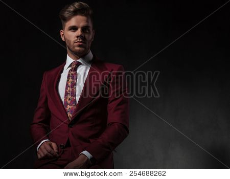 portrait of handsome stylish man in red suit sitting on grey wallpaper background and looking to side