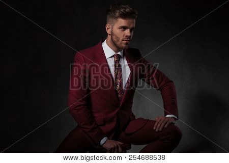 portrait of elegant man wearing a grena suit looking to side while sitting on a wooden chair on grey wallpaper background