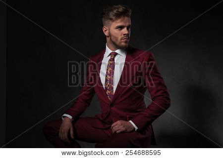 man wearing a grena suit looks to side while sitting on a wooden chair on grey wallpaper background
