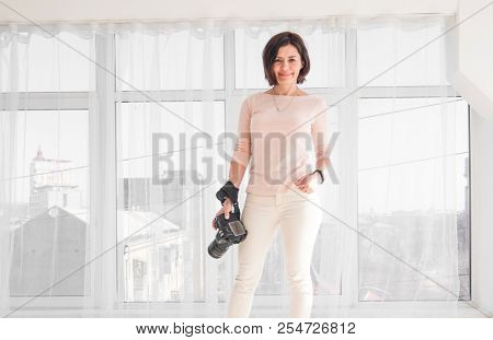 Attractive woman with dslr camera in hands smiling and posing in light white room with big window. Happy smiling female photographer in front of bright windows