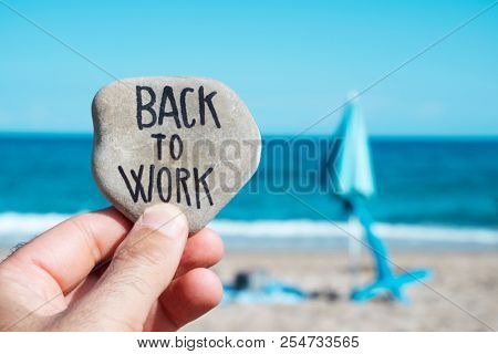closeup of the hand of a young caucasian man on the beach, in front of a folded beach umbrella and the ocean in the background, holding a stone with the text back to work written in it