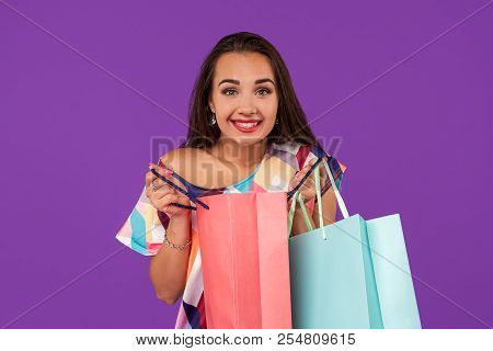 Beautiful Woman Peeks Into A Package, In The Hands Of Multi-colored Shopping Bags On A Purple Backgr