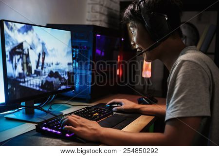 Asian cheerful gamer boy playing video games online on computer in dark room wearing headphones with microphone and using backlit colorful keyboard