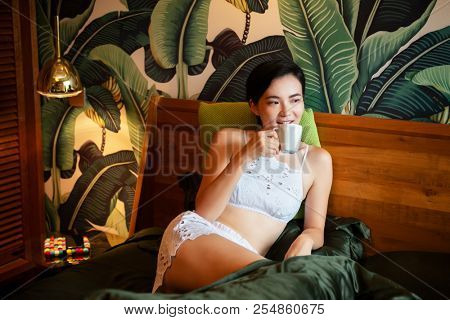 Beautiful chinese young woman wearing pajamas lies in bed and drinks coffee. Lazy morning. Happy sleppy girl wit cup og tea in bedroom.