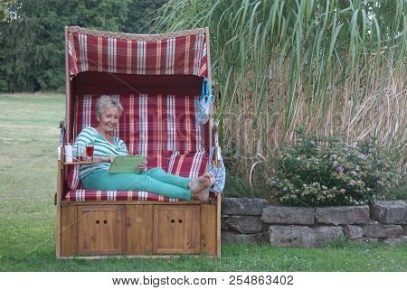 In The Roofed Wicker Beach Chair Reading Funny Emails On The Tablet, The Attractive Woman Is Happy A