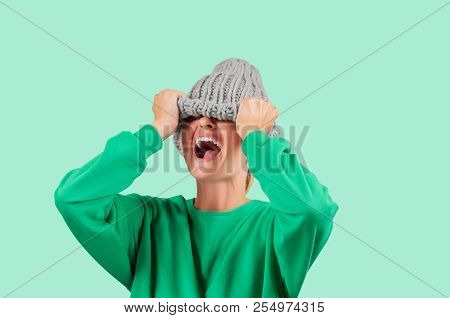 Furious. Emotional Angry Woman Is Pulling Wool Hat Over Her Eyes On Green Background
