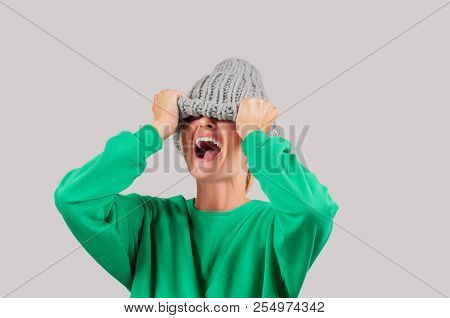Furious. Emotional Angry Woman Is Pulling Wool Hat Over Her Eyes On Gray Background