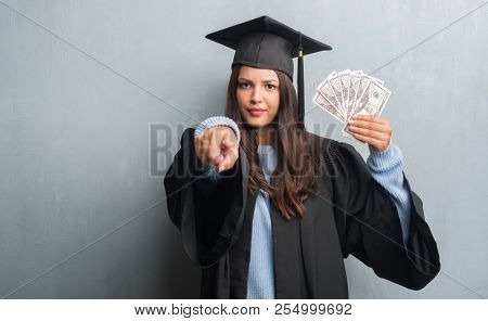 Young brunette woman over grunge grey wall wearing graduate uniform holding dollars pointing with finger to the camera and to you, hand sign, positive and confident gesture from the front