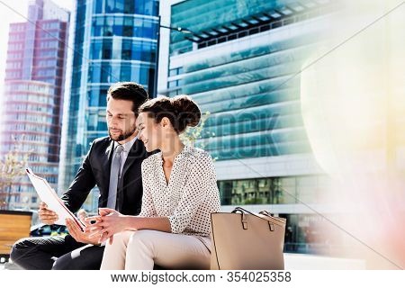 Young attractive businessman discussing business plans over document with beautiful businesswoman while sitting on bench against office building