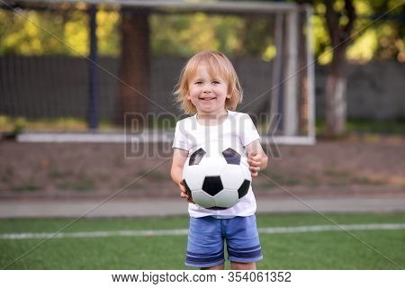Happily Smiling Blonde Toddler Boy Standing In Football Field (stadium) With Soccer Ball Against Goa