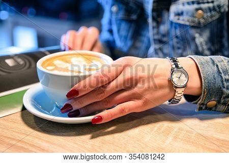 Close Up View On Red Gel Nails Of Girl Holding Cup Of Cappucinno And Reading Magazine In Cafe